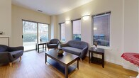 This single-floor space is designed as an open-plan communal area that seamlessly integrates a dining zone and a lounge area under one cohesive layout. The warm, light-toned hardwood flooring runs continuously throughout, unifying both functional zones and contributing to the space's welcoming ambiance.

The dining area is anchored by a large rectangular table accommodating up to eight guests, surrounded by dark-wood ladder-back chairs with upholstered seats. A pendant light fixture hangs overhead, complemented by wall-mounted sconces that cast a soft, warm glow. A built-in counter runs along one wall, offering additional utility, while a vibrant floral artwork adds a decorative accent. Multiple windows along the dining perimeter invite views of surrounding greenery and fill the area with natural daylight.

The lounge area features a compact sofa and a rounded accent chair, both upholstered in charcoal-grey fabric and arranged around a dark coffee table. Additional side tables provide functional surfaces. A bank of large sliding and fixed windows lines the exterior wall, allowing abundant natural light and views to the outdoors. Wall sconces provide supplemental lighting in the evening.

The textured ceiling, neutral warm-beige walls, and consistent flooring throughout give the space a clean, modern, and inviting character well-suited for social gatherings and shared use.