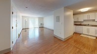 Located at 27 Page Ter in Stoughton, Massachusetts, this single-floor unit offers a thoughtfully laid-out interior with a clean, neutral aesthetic throughout. The expansive open-concept living and dining area is anchored by warm hardwood-style flooring that reflects natural light, a large sliding glass door with vertical blinds, and a wall-mounted electrical panel that adds a practical touch. Adjacent to the living space, the kitchen features white cabinetry with sleek bar-pull hardware, taupe laminate countertops, a stainless steel sink, and white appliances including a dishwasher and range — all set above matching hardwood-style flooring. A closeted storage area and additional open space complete the main living zone. The bedrooms are generously sized, each finished with plush neutral carpeting, large windows offering leafy outdoor views, wall-unit HVAC systems, and baseboard heating. The unit includes two full bathrooms: one spacious bath with a tub-shower combination and light tile flooring, and a second bath featuring a modern white vanity with a large mirror, updated chrome fixtures, and a shower. Natural light flows freely throughout, creating a bright and welcoming ambiance across every room.