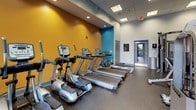 This fitness center is designed to support a comprehensive workout experience within a single, open-plan floor. The space is visually energized by bold accent walls in teal and amber-orange, contrasting with neutral gray tones throughout the rest of the room. The flooring consists of durable rubber matting, practical for both strength and cardio areas.

The strength training zone is well-stocked, featuring a full rack of dumbbells in varying weights, kettlebells, Nautilus and Hoist weight machines, a cable pulley system, adjustable benches, and an assortment of exercise balls. Large wall-to-wall mirrors along one side visually expand the space and allow users to monitor their form during workouts.

The cardio section lines another wall and includes multiple Matrix treadmills, elliptical trainers, and upright bikes, all facing large floor-to-ceiling windows and glass doors that open to an outdoor area, providing natural light and exterior views during exercise.

Overhead, a drop ceiling with recessed and panel lighting ensures the space is evenly and brightly lit. The layout is open and organized, allowing for fluid movement between equipment zones. The overall ambiance is modern, functional, and motivating.