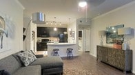 This single-floor residence showcases an open-concept layout that connects the living and kitchen areas into one cohesive, functional space. The kitchen is outfitted with dark espresso cabinetry, black appliances including a refrigerator and range, and light-toned countertops that provide a striking contrast. A peninsula-style breakfast bar with seating for two serves as both a dining and entertaining hub, anchored by pendant lighting above. Track lighting along the kitchen ceiling ensures excellent task illumination throughout the cooking area. The living area flows naturally from the kitchen, featuring hardwood-style flooring that runs continuously across the space, lending warmth and visual continuity. Crown molding along the upper walls adds a refined architectural detail. Neutral, warm wall tones keep the interior feeling bright and welcoming. A hallway visible from the main living area leads to a bathroom, suggesting a thoughtfully arranged floor plan. The overall ambiance is contemporary and practical, balancing style with everyday functionality through cohesive finishes and an efficient, well-lit open design.