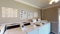 This shared laundry facility is a single-story space designed to provide residents with convenient, on-site laundry services. The room is finished with neutral gray walls accented by a white chair-rail border, creating a clean and utilitarian aesthetic. Dark hardwood-style flooring runs throughout, adding a degree of visual warmth to the functional space.

The facility houses multiple commercial-grade top-loading washing machines lining one wall, complemented by stacked washer-dryer units positioned along another section of the room. Instructional signage is posted prominently on the walls, offering guidance on machine operation in both English and Spanish, ensuring accessibility for all users.

A fold-out utility table provides a convenient surface for sorting and folding laundry, and a wicker basket is available for resident use. A ceiling light fixture provides adequate overhead illumination throughout the space.

A full-glass entry door opens directly to an outdoor walkway and landscaped grounds, allowing natural light to filter into the room and providing easy access from surrounding buildings. A fire safety box is mounted near the entrance, reflecting standard safety compliance. Overall, the facility is straightforward and practical, focused on delivering an efficient laundry experience.