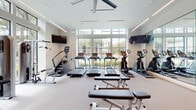 This contemporary fitness center is thoughtfully designed to support a comprehensive workout experience. The space is anchored by a bright, open cardio zone outfitted with multiple treadmills, elliptical machines, stair climbers, and rowing machines, all arranged to face expansive floor-to-ceiling windows that frame lush outdoor views. The generous natural light flowing through the large black-framed windows creates an energizing atmosphere throughout the facility. A full-wall mirror runs the length of one side of the main exercise area, visually expanding the space and allowing users to monitor their form during workouts. Ceiling fans and linear recessed lighting complement the clean, neutral color palette of white walls and light carpet flooring. A dedicated strength and functional training zone accommodates cable machines, a multi-functional weight station, free weights on a rack, and flat benches, providing ample options for resistance training. An adjacent area includes medicine balls, stability balls, and a flat-screen television, offering space for stretching and functional fitness. A small wellness station with supplies and shelving is neatly tucked into one corner. Multiple wall-mounted televisions are positioned throughout the space for entertainment during workouts.