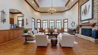 This generously sized single-floor community space is designed with an upscale, traditional aesthetic that blends warmth and functionality throughout. The architecture is immediately distinguished by dramatically high ceilings accented with rich wood beam detailing and a tiered tray ceiling in the main lounge area, where a crystal chandelier serves as a striking focal point.

The primary lounge area features rich hardwood flooring, wood-trimmed windows lining multiple walls, and a large stone fireplace framed by decorative artwork. Seating arrangements accommodate both intimate conversation and larger gatherings, creating a welcoming, versatile atmosphere.

An adjacent dining area offers a substantial dark-finished conference-style table surrounded by upholstered chairs, ideal for meetings or group dining, bathed in natural light from tall wood-framed windows with outdoor views.

The entryway or foyer transitions between spaces using large-format tile flooring, exposed wood beam doorframes, and display cabinetry, offering clear sightlines throughout. A well-equipped kitchen features rich wood cabinetry, granite countertops, stainless steel appliances, and a secondary galley-style prep kitchen, supporting catering and event needs efficiently.

Overall, the space conveys an elegant yet approachable character suited for community gatherings and events.