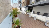 This single-level outdoor space is a thoughtfully designed urban deck that blends modern aesthetics with a relaxed, lounge-like atmosphere. The warm-toned composite wood decking spans the length of the space, providing a clean and durable foundation that anchors the overall design. A continuous built-in bench lines one side of the deck, dressed with black-and-white striped and solid cushions and pillows, offering generous seating capacity ideal for social gatherings or events.

White hourglass-shaped accent stools are distributed across the deck, complementing the modern, minimalist furnishing style. Large white planters house tropical palms and agave plants, introducing vibrant greenery that contrasts beautifully against the whitewashed stucco walls and natural concrete block boundary wall. A decorative oversized silver sphere planter adds a sculptural focal point to the seating area.

String lights are strung overhead across the width of the deck, creating an inviting ambiance that transitions seamlessly from day to evening use. The space is open to the sky, allowing abundant natural light during the day. Surrounding multi-story buildings with balconies frame the courtyard, lending an urban character. Black metal railings and a concrete pathway area provide structured access points, completing this well-appointed outdoor venue.