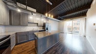 Located at 515 St Germain St W in Saint Cloud, MN, this four-bedroom, two-bathroom unit offers a distinctive urban loft aesthetic across a single floor. The space is defined by its soaring ceilings with exposed ductwork, dark-stained ceiling beams, and rich hardwood flooring throughout the main living and kitchen areas. The kitchen serves as a centerpiece, featuring gray shaker-style cabinetry, granite countertops, a large central island with pendant lighting, stainless steel appliances including a French door refrigerator, and track lighting overhead. A glass sliding door from the kitchen and living area opens to an outdoor balcony, allowing natural light to fill the space. The four bedrooms are carpeted for comfort and are connected via hallways that maintain the loft's clean, modern palette of light gray walls and white trim. Closets provide dedicated storage within the bedroom areas. The two bathrooms are finished with tile flooring, dark wood vanities, white countertops, and brushed nickel hardware — one featuring a full tub-shower combination. The overall ambiance blends industrial character with contemporary finishes, creating a functional and visually striking living environment.