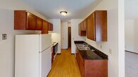 This single-floor residence offers a thoughtfully laid-out living environment across several distinct functional areas. The living room is open and airy, featuring soft carpeted flooring, neutral walls, a textured ceiling, and a large sliding glass door that floods the space with natural light while providing access to an outdoor balcony with exterior views. A baseboard heating system runs along the perimeter, ensuring comfort throughout.

The galley-style kitchen is efficiently designed with rich, warm-toned cabinetry extending to the upper walls for generous storage, dark granite countertops, a double stainless steel sink, and durable hardwood-style flooring. White appliances including a refrigerator and range complete the functional setup.

The bedroom features plush carpeting, a window dressed with vertical blinds that allow natural light, a textured ceiling with a flush-mount light fixture, and baseboard heating. A separate room showcases an expansive wall of built-in closets with panel doors, offering exceptional storage capacity alongside additional carpeted living space.

The full bathroom is clean and straightforward, with tile flooring, a combined tub and shower unit, a toilet, and a single vanity with mirror. Dark wood-toned doors add a classic contrast throughout the unit. Overall, the space presents a functional, comfortable layout with consistent neutral finishes.