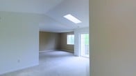This single-floor space showcases a thoughtfully designed open layout bathed in natural light. A centrally positioned skylight draws the eye upward to the vaulted ceiling, flooding the interior with daylight and lending an airy, expansive feel to the room. The ceiling's angled architecture adds visual interest and a sense of height beyond what standard construction typically provides.

The walls are finished in warm, neutral tan tones that complement the soft, plush carpeting underfoot, creating a cohesive and inviting atmosphere throughout. White trim along the baseboards provides clean contrast against both the wall color and flooring.

A large multi-pane window along the back wall invites additional natural light while offering views of the surrounding greenery. Adjacent to it, a full-length glass door provides direct access to an outdoor deck or patio area, further connecting the interior to the outdoors and enhancing the sense of openness.

The space is currently unfinished in terms of furnishings, making it highly adaptable for a variety of uses. The neutral palette and abundant natural light make it a welcoming, move-in-ready environment suitable for a range of configurations and personal styles.