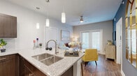 This thoughtfully designed 1-bedroom, 1-bathroom residence offers a cohesive open-concept floor plan that maximizes every square foot. The living and kitchen areas flow seamlessly together, anchored by rich hardwood-style flooring that extends throughout the unit. The kitchen is a standout feature, showcasing dark espresso cabinetry, granite countertops, stainless steel appliances including a refrigerator, range, and microwave, along with a peninsula island with a deep double-basin sink and elegant pendant lighting overhead. A striking blue accent wall with arched decorative mirrors adds a bold architectural focal point between the kitchen and hallway. The living area is brightened by large windows and a glass sliding door that invites natural light, complemented by a ceiling fan above. The bedroom is a serene retreat, featuring hardwood-style flooring, generous natural light from a large window, and a sliding barn door that reveals a neatly tucked stacked washer and dryer unit — a rare and practical convenience. The bathroom is finished with wood-toned cabinetry, granite countertops, tile flooring, and a glass-enclosed walk-in shower. A dedicated closet completes the layout, providing ample storage throughout this efficiently designed home.