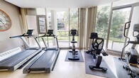 Located at 22 Elkhorn Dr in Toronto's North York neighbourhood, this exercise room offers a well-equipped fitness space bathed in natural light. Floor-to-ceiling windows and glass sliding doors line the exterior wall, providing expansive views of the surrounding greenery and adjacent building grounds, creating an open and inviting atmosphere for workouts.

The room features polished terrazzo flooring throughout, lending a clean and durable surface well-suited for a fitness environment. A ceiling fan is centrally mounted, complementing a wall-mounted air conditioning unit to ensure comfortable year-round use. Recessed and wall-mounted light fixtures provide ample illumination throughout the space.

The exercise room is equipped with a variety of cardio machines, including two treadmills and an elliptical trainer, as well as a stationary bike, all positioned to take advantage of the natural light and outdoor views. A multi-tier dumbbell rack stocked with a range of free weights occupies one corner, catering to strength training needs. A decorative wall clock adds a functional and stylistic accent to the neutral-toned walls.

The room is accessible via a glass-panelled door, and baseboard heating along the interior wall ensures warmth during cooler months. Overall, this fitness space is practical, bright, and thoughtfully arranged for an effective workout experience.