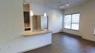 This single-floor space showcases a thoughtfully designed open-concept layout that seamlessly connects the kitchen and living areas. The kitchen is highlighted by a peninsula-style breakfast bar topped with granite countertops and an integrated stainless steel sink, making it both a functional workspace and a social hub. Elegant pendant lights hang above the bar, complementing the track lighting system installed on the ceiling, which provides ample illumination throughout the space. Dark-toned cabinetry in the kitchen offers a striking contrast against the neutral walls, while open shelving adds a practical and modern touch. The living area benefits from natural light streaming through a large window fitted with blinds, creating a bright and welcoming atmosphere. Rich hardwood-style flooring runs continuously across the entire space, tying the design together with warmth and consistency. The clean lines, neutral color palette, and contemporary fixtures combine to give the space a modern, move-in-ready aesthetic that is both stylish and highly functional.