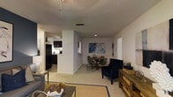 This single-floor residence is designed with an open-concept floor plan that seamlessly connects the primary living areas, creating a cohesive and functional layout. The living room serves as the heart of the space, featuring plush carpeted flooring, a woven area rug, and an assortment of comfortable seating including a navy blue accent chair with nailhead trim. A rustic wooden media console adds a warm, textured contrast to the neutral walls.

Adjacent to the living room, the dining area provides an intimate setting with a round wooden table surrounded by sleek metal-framed chairs. A large abstract canvas artwork in cool blue tones anchors the dining space, adding a contemporary artistic touch to the room.

The kitchen is separated by a partial wall, offering both openness and definition. It is equipped with dark cabinetry, a black refrigerator, and a light-colored countertop, creating a bold contrast. A track lighting fixture illuminates the workspace efficiently.

Throughout the space, recessed lighting and ceiling vents contribute to a clean, finished look. The textured ceiling adds architectural character, while the neutral wall tones provide a versatile backdrop that complements a variety of décor styles. Overall, the space delivers a balanced blend of practicality and modern comfort.