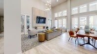This single-floor community clubhouse showcases a thoughtfully designed open-concept layout that seamlessly blends comfort and sophistication. Soaring double-height ceilings and an expansive wall of floor-to-ceiling windows flood the interior with natural light, creating an airy, welcoming atmosphere throughout.

The main lounge area is anchored by a striking floor-to-ceiling wood-paneled fireplace surround with a linear marble hearth, framed by a wall-mounted flat-screen TV. A sculptural globe chandelier serves as a focal point overhead, while plush sectional seating, accent chairs, and layered area rugs define an inviting gathering space. Adjacent to the lounge, a wooden communal dining table with tan leather chairs provides a relaxed setting for socializing.

The social kitchen area is equally impressive, featuring a large waterfall-edge marble island with bar seating in navy upholstered chairs, pendant lighting with gold accents, and a dramatic forest-motif feature wall as a backdrop. Stainless steel appliances, including a full-size refrigerator, complete the space.

The entry corridor is bright and gallery-like, adorned with colorful framed artwork, a mid-century modern credenza, and a grid of clerestory windows that draw the eye upward. Light wood-look flooring runs throughout, tying each zone together cohesively and reinforcing the space's clean, contemporary aesthetic.
