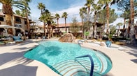 This expansive resort-style outdoor amenity area is thoughtfully designed for relaxation and entertainment. At its centerpiece sits a freeform swimming pool with crystal-clear water and a dramatic natural rock waterfall feature, evoking a tropical oasis atmosphere. Towering palm trees frame the pool deck, providing shade and a lush, resort-like ambiance. Adjacent to the pool, a separate spa offers a dedicated space for unwinding, surrounded by comfortable poolside lounge furniture.

The covered patio area is a standout feature, supported by elegant columned architecture with a stucco finish in a warm desert palette. Ceiling fans are installed throughout the covered space, ensuring comfort in warmer months. Modern outdoor sofas and chairs with crisp white cushions are arranged for casual gatherings, while a sliding glass door provides seamless indoor-outdoor access.

A dedicated outdoor lounge area features wicker seating with plush white cushions centered around a fire pit table, shaded by a large striped umbrella — ideal for intimate gatherings. The grilling station is equipped with dual stainless steel built-in gas grills, making it well-suited for outdoor entertaining. Decorative ironwork gates, manicured landscaping with artificial turf accents, and desert-adapted plants complete the polished, cohesive aesthetic of this amenity space.