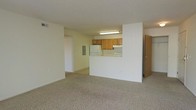This single-floor unit offers a practical and functional layout designed for comfortable everyday living. The spacious living area is finished with wall-to-wall carpet flooring and features large sliding glass doors dressed with vertical blinds, allowing abundant natural light to fill the room while providing access to an outdoor area with exterior views.

The kitchen is positioned at the heart of the unit, separated from the living space by a half-wall partition that creates a semi-open feel while maintaining a defined cooking area. The kitchen is equipped with warm-toned oak cabinetry, a white refrigerator, and a range, all illuminated by overhead fluorescent lighting. An electrical panel is conveniently located on the kitchen-adjacent wall.

Neutral, light-toned walls run throughout the unit, contributing to a bright and airy atmosphere. A hallway visible from the main living area leads to additional rooms, suggesting a well-organized floor plan. The space also includes a door to what appears to be a closet or utility area, adding to the overall storage options. The clean, simple design provides a versatile canvas suitable for a variety of living arrangements.