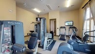 This fitness center offers a thoughtfully organized workout environment spread across a single level. The space is finished with warm, neutral-toned walls and carpeted flooring accented by rubber exercise mats positioned beneath the equipment for added safety and stability. Fluorescent lighting ensures consistent illumination throughout, while multiple wall-mounted flat-screen televisions are strategically placed to keep users entertained during workouts.

The cardio section is well-appointed with multiple treadmills, elliptical cross-trainers, and a stationary cycle, providing a broad range of aerobic options. A dedicated strength-training area features a multi-station cable machine, a leg press unit, adjustable benches, and free weights, catering to users focused on resistance and muscle conditioning.

A standout feature of the space is the row of tall, elegantly draped windows along one wall, which flood the room with natural light and offer pleasant views of the surrounding greenery. The neutral drapery adds a refined aesthetic touch uncommon in typical fitness facilities. An adjacent doorway suggests access to a restroom or utility area, adding practical convenience. Overall, the fitness center balances functionality with a comfortable, well-maintained ambiance.