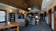 This single-floor rustic retreat blends natural materials and warm design elements to create an inviting, cabin-like atmosphere. The centerpiece of the open-concept living area is a commanding floor-to-ceiling stone fireplace, adding both visual drama and a sense of rugged charm. Flanking the fireplace, built-in bookshelves offer both functionality and character.

The vaulted ceiling, clad in rich wood planks and accented by dual ceiling fans, draws the eye upward and amplifies the sense of spaciousness throughout the main living space. Recessed lighting complements the warm overhead pendants, ensuring the interior remains well-lit at all times.

The living area is furnished with plush leather sofas and accent tables, creating a comfortable gathering space centered around the hearth. Adjacent to the living room, a dining area with a wooden table provides a casual setting for meals.

The kitchen flows seamlessly from the living space in an open-concept layout. It features dark cabinetry, stainless steel appliances including a refrigerator, and ample counter space, making it well-suited for full-time living or extended stays.

The space is finished with deep charcoal carpeting that grounds the warm wood and stone tones throughout. A solid wood entry door adds to the overall sense of craftsmanship and rustic elegance that defines this distinctive space.