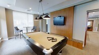 This community lounge is designed to offer residents a comfortable and engaging social space. At the heart of the room sits a full-size pool table with a cream felt surface, illuminated by a stylish industrial-style pendant light fixture with three hanging shades — a standout design element that anchors the space. Opposite the table, a warm wood accent wall provides a rich, natural backdrop for a wall-mounted flat-screen TV, creating an ideal setup for both gaming and entertainment. The room benefits from generous natural light streaming through large windows dressed with light-filtering roller shades, giving the space an open and airy feel. A seating area near the windows offers a quieter retreat, furnished with a round table and upholstered chairs — perfect for casual conversation or relaxing between games. The flooring features a neutral carpet that complements the room's contemporary yet inviting aesthetic. Recessed lighting supplements the natural light, ensuring the space is well-lit throughout the day and into the evening. Overall, the community lounge strikes a balance between recreational functionality and relaxed comfort, making it a versatile gathering place for residents.