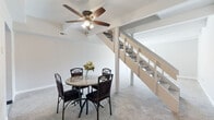 This two-floor space is designed with a clean, neutral aesthetic that carries consistently throughout. The main level features an open layout anchored by a dining area with plush carpeting and a ceiling fan overhead. A striking open staircase with a painted wood railing serves as a central architectural element, connecting the two floors while contributing to a sense of openness. Adjacent to the dining area, the galley-style kitchen is thoughtfully updated with light gray shaker cabinets, matte black hardware, a textured tile backsplash, a stainless steel sink, and wood-toned laminate flooring. A window above the sink brings in natural light and views of surrounding greenery. A hallway near the base of the stairs provides access to a carpeted lower-level room and features a classic six-panel door. The upper floor includes generously sized carpeted rooms finished in soft gray tones, each with windows that admit natural light. A full bathroom on this level is finished with wood-look tile flooring, a white vanity with ample drawer storage, matte black fixtures, and a tub-shower combination. Throughout both floors, white trim and neutral wall tones create a cohesive, move-in-ready atmosphere.
