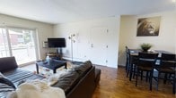 This 2-bedroom, 2-bathroom single-floor residence offers a straightforward and functional layout designed for comfortable everyday living. The open-plan living and dining area serves as the heart of the home, anchored by warm hardwood flooring that runs throughout the space. The living area accommodates a full-sized sofa and coffee table arrangement, complemented by a wall-mounted television and a stylish floor lamp, creating a relaxed and inviting atmosphere. A wall-unit air conditioner and baseboard heating ensure year-round climate comfort. Adjacent to the living space, a dining area features a raised table with bar-style seating, making it ideal for casual meals or a workspace. The white-painted walls and standard ceiling height give the space a clean, neutral backdrop that is easy to personalize. The entry door opens directly into the main living area, providing convenient access. With its classic hardwood floors, practical amenities, and an efficiently organized open-concept common area, this residence delivers a no-frills yet comfortable living environment suited for a range of occupants.