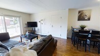 This 2-bedroom, 2-bathroom single-floor residence offers a straightforward and functional layout designed for comfortable everyday living. The open-plan living and dining area serves as the heart of the home, anchored by warm hardwood flooring that runs throughout the space. The living area accommodates a full-sized sofa and coffee table arrangement, complemented by a wall-mounted television and a stylish floor lamp, creating a relaxed and inviting atmosphere. A wall-unit air conditioner and baseboard heating ensure year-round climate comfort. Adjacent to the living space, a dining area features a raised table with bar-style seating, making it ideal for casual meals or a workspace. The white-painted walls and standard ceiling height give the space a clean, neutral backdrop that is easy to personalize. The entry door opens directly into the main living area, providing convenient access. With its classic hardwood floors, practical amenities, and an efficiently organized open-concept common area, this residence delivers a no-frills yet comfortable living environment suited for a range of occupants.
