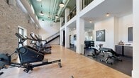 Located at 1720 Nick Nuccio Parkway in Tampa, FL, this two-story fitness facility offers a well-appointed and architecturally distinctive workout environment. The ground floor greets visitors with soaring double-height ceilings, exposed brick walls, and an industrial-chic aesthetic highlighted by pendant lighting and large windows that invite abundant natural light. The flooring transitions between polished concrete in the free weights area and warm cork flooring in the cardio and functional training zones, providing both durability and comfort underfoot. A comprehensive dumbbell rack, adjustable benches, and a full array of strength equipment, including power racks and cable machines, make this level suitable for a wide range of strength training routines. StairMaster rowers and additional cardio machines are also thoughtfully arranged throughout. A sleek black metal staircase connects to the second floor, which continues the facility's refined design with floor-to-ceiling mirrors, additional cable and functional training machines, stationary bikes, and open floor space ideal for stretching or auxiliary exercises. The upper level also features an iron-railed mezzanine overlooking the main floor, enhancing the sense of openness. Together, both floors deliver a comprehensive, modern fitness experience with a strong architectural character.