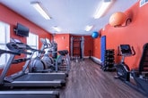 Located at 3300 N Ingle Pl in Macon, Georgia, this fitness center is a well-equipped, single-floor facility designed to support a comprehensive workout routine. The space is immediately defined by its bold burnt-orange walls, which create an energizing atmosphere complemented by durable wood-look flooring throughout.

The cardio zone features multiple treadmills, elliptical machines, and a stationary bike, with a wall-mounted television providing entertainment during workouts. Natural light filters in through windows fitted with blinds, while overhead fluorescent lighting ensures the space remains bright and functional.

A large wall mirror spans one side of the room, visually expanding the space and allowing users to monitor their form during exercise. The strength training area includes a multi-functional cable machine with an adjustable bench, ideal for a wide range of resistance exercises. Additional equipment includes a dumbbell rack with a full set of weights, medicine balls, stability balls in multiple sizes, a yoga mat, and battle ropes for high-intensity interval training.

A dedicated signage board near the entrance outlines fitness center hours and community guidelines, reinforcing the space's role as a shared amenity. The layout is organized and open, allowing for smooth traffic flow between equipment stations and accommodating a variety of fitness disciplines in one convenient location.