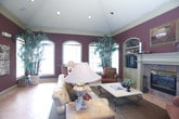 This single-story space showcases a warm, traditional aesthetic defined by deep burgundy walls, decorative white pilasters, and crown molding that add architectural character throughout. The main living area is generously proportioned, anchored by a classic fireplace with a detailed white mantel and stone surround, set beneath soaring vaulted ceilings fitted with recessed lighting. Large arched windows allow natural light to filter in, enhancing the richness of the warm color palette.

The living area features tiled flooring in a neutral earth tone, complemented by area rugs that define seating zones. A tufted leather sofa and upholstered accent chairs create a refined, comfortable arrangement centered around a stone-top coffee table. Built-in shelving flanks the fireplace, providing display space.

An arched opening connects the living space to a kitchen area, which features a raised breakfast bar with barstool seating, white cabinetry, and pendant lighting overhead. A separate entry area includes a console table beneath framed artwork and leads to a bright adjoining room through white louvered French doors. Decorative wallpaper accents, framed coastal artwork, and layered lighting throughout contribute to the space's cohesive and inviting traditional ambiance.