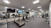 Located at 3101 Euclid Ave in Cleveland, Ohio, this fitness center offers a well-organized, single-floor workout facility designed to accommodate a comprehensive range of exercise routines. The space features a clean, modern aesthetic with neutral grey walls, a drop-panel ceiling equipped with recessed fluorescent lighting, and durable commercial-grade carpet flooring in a textured grey tone throughout.

The cardio section is prominently arranged along one side of the room and includes multiple treadmills, elliptical machines, and a stationary bike, all by Spirit. Wall-mounted flat-screen televisions are positioned above the cardio equipment, providing entertainment during workouts. Full-length mirrors line an entire wall, visually expanding the space and allowing users to monitor their form.

A dedicated strength training area features a multi-functional cable machine and a rack stocked with a full range of dumbbells, offering options for both beginner and advanced weightlifting. Additional equipment includes a rowing machine, a weight bench, and an assortment of stability balls and medicine balls, supporting functional and cross-training workouts.

The facility is accessed through a solid double-door entry with windows and clearly marked emergency exits, ensuring safety compliance. The overall ambiance is clean, professional, and purposefully functional, making this fitness center a well-equipped space for daily exercise.