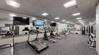 Located at 3101 Euclid Ave in Cleveland, Ohio, this fitness center offers a well-organized, single-floor workout facility designed to accommodate a comprehensive range of exercise routines. The space features a clean, modern aesthetic with neutral grey walls, a drop-panel ceiling equipped with recessed fluorescent lighting, and durable commercial-grade carpet flooring in a textured grey tone throughout.

The cardio section is prominently arranged along one side of the room and includes multiple treadmills, elliptical machines, and a stationary bike, all by Spirit. Wall-mounted flat-screen televisions are positioned above the cardio equipment, providing entertainment during workouts. Full-length mirrors line an entire wall, visually expanding the space and allowing users to monitor their form.

A dedicated strength training area features a multi-functional cable machine and a rack stocked with a full range of dumbbells, offering options for both beginner and advanced weightlifting. Additional equipment includes a rowing machine, a weight bench, and an assortment of stability balls and medicine balls, supporting functional and cross-training workouts.

The facility is accessed through a solid double-door entry with windows and clearly marked emergency exits, ensuring safety compliance. The overall ambiance is clean, professional, and purposefully functional, making this fitness center a well-equipped space for daily exercise.
