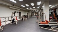 This fitness center offers a thoroughly equipped, single-floor exercise facility designed to support a wide range of workout routines. The space is defined by durable speckled rubber flooring throughout, providing both safety and comfort underfoot. Overhead, a grid of recessed and panel lighting ensures the entire area is consistently and brightly illuminated.

The strength training zone is generously outfitted with a variety of selectorized cable and weight machines featuring distinctive red-accented upholstery, along with a dumbbell rack stocked with a full range of free weights. A dedicated raised platform area, accessible via a short staircase with handrails, houses cardiovascular equipment including treadmills and an elliptical trainer. Bold orange accent walls punctuate this section, adding visual energy to the space.

Structural columns are integrated throughout the floor plan and the facility benefits from clear sightlines across the main workout area. A set of large glass-paneled double doors connects the fitness center to an adjoining common area, enhancing the sense of openness. Multiple exit points are clearly marked, and the layout allows for comfortable circulation between equipment. Overall, the space presents a clean, functional, and motivating environment suited for a broad range of fitness activities.