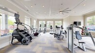 Located at 665 Lafayette Ave in Fayetteville, Georgia, this fitness center offers a comprehensive and thoughtfully designed exercise environment across a single open floor. The space features a sophisticated coffered ceiling with recessed lighting and a large ceiling fan, ensuring ample air circulation and a polished aesthetic throughout. Neutral-toned walls and herringbone-patterned carpet tiles contribute to a clean, modern ambiance.

Natural light floods the room through multiple large windows lining the perimeter walls, providing views of the surrounding greenery and creating a welcoming atmosphere for workouts. Full-length wall mirrors in the strength training area further enhance the sense of openness and allow users to monitor their form.

The cardio section is extensively equipped with treadmills, elliptical machines, stair climbers, and stationary cycling bikes — many featuring individual digital screens. The strength training area includes a multi-station cable machine, a Smith machine with a weight bench, selectorized resistance machines, and a dedicated free weights zone with a dumbbell rack and medicine ball storage. A wall-mounted television adds an entertainment option for users. The well-organized layout maximizes the available floor space, making the fitness center suitable for a wide variety of exercise routines and fitness levels.