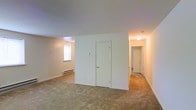 This single-floor space offers a straightforward and functional layout designed for comfortable everyday living. The main living area is generously proportioned, featuring wall-to-wall carpeting that adds warmth underfoot and neutral white walls that create a clean, bright backdrop throughout. A central partition wall serves as a defining architectural element, incorporating a closet with a white paneled door accented by a brass knob, effectively dividing the open space while maintaining a sense of flow. A window on the adjacent wall allows natural light to filter into the living area, complementing the baseboard heating unit positioned beneath it. A hallway extends from the main living space, connecting to additional rooms toward the rear of the unit, including what appears to be a bathroom and further living or sleeping quarters. The hallway is illuminated by a ceiling-mounted light fixture, ensuring consistent visibility throughout. The overall color palette remains soft and neutral, with white ceilings and walls pairing seamlessly with the tan carpeting. The layout is compact yet well-organized, making efficient use of the available square footage across its single floor.