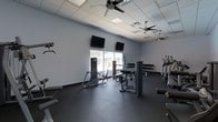 This fitness center offers a well-organized and fully equipped workout environment designed to accommodate a range of exercise routines. The space features a clean, modern aesthetic with light gray walls and a drop-panel ceiling outfitted with multiple ceiling fans, ensuring consistent airflow throughout the room. Dark rubberized flooring covers the workout area, providing both durability and safety underfoot.

The strength training section is prominently featured, housing a selection of selectorized weight machines, including what appears to be a leg press and a seated cable or chest machine. A full dumbbell rack lines one wall, offering a wide range of free weight options for varied training needs. On the opposite side, a row of cardio machines — including treadmills — is neatly arranged, making efficient use of the available floor space.

Two wall-mounted flat-screen televisions are positioned for easy viewing from multiple workout stations, adding an entertainment element to training sessions. A window along the back wall allows natural light to filter into the space, creating a bright and open atmosphere. The overall layout is spacious and uncluttered, allowing for comfortable movement between equipment. This fitness center provides a functional and inviting environment suitable for both strength and cardiovascular training.