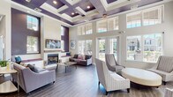 Located at 7521 Windmill Harbor Way in Raleigh, NC, this sophisticated single-story clubhouse is designed to impress at every turn. The main lounge serves as the centerpiece of the space, boasting dramatically high ceilings adorned with a striking coffered design painted in deep purple and white — a bold architectural statement that sets the tone throughout. Wood-look tile flooring runs continuously across the entire space, lending warmth and cohesion to every area.

A floor-to-ceiling fireplace anchors one end of the lounge, framed by built-in window bench seating with upholstered cushions, creating a cozy gathering spot. A wall-mounted television above the mantel adds a modern touch. Large French doors and expansive windows flood the room with natural light and offer direct views of the outdoor pool area, seamlessly connecting the interior to the exterior.

Adjacent to the main lounge is a versatile secondary seating area, complete with its own wall-mounted TV and comfortable furnishings — ideal for casual lounging or small group gatherings. A dedicated workspace area with workstations provides a functional environment for productivity. A dining nook with a round table and modern chairs offers additional flexibility for meetings or informal dining.

The overall ambiance is polished yet approachable, with a cohesive palette of soft grays, whites, and rich purple accents throughout.