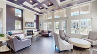 Located at 7521 Windmill Harbor Way in Raleigh, NC, this sophisticated single-story clubhouse is designed to impress at every turn. The main lounge serves as the centerpiece of the space, boasting dramatically high ceilings adorned with a striking coffered design painted in deep purple and white — a bold architectural statement that sets the tone throughout. Wood-look tile flooring runs continuously across the entire space, lending warmth and cohesion to every area.

A floor-to-ceiling fireplace anchors one end of the lounge, framed by built-in window bench seating with upholstered cushions, creating a cozy gathering spot. A wall-mounted television above the mantel adds a modern touch. Large French doors and expansive windows flood the room with natural light and offer direct views of the outdoor pool area, seamlessly connecting the interior to the exterior.

Adjacent to the main lounge is a versatile secondary seating area, complete with its own wall-mounted TV and comfortable furnishings — ideal for casual lounging or small group gatherings. A dedicated workspace area with workstations provides a functional environment for productivity. A dining nook with a round table and modern chairs offers additional flexibility for meetings or informal dining.

The overall ambiance is polished yet approachable, with a cohesive palette of soft grays, whites, and rich purple accents throughout.