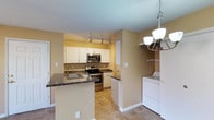 This single-floor residence offers a functional and well-appointed layout with a focus on practical living. The kitchen serves as the heart of the home, showcasing white cabinetry that extends to the ceiling, dark granite-style countertops, and a full suite of stainless steel appliances including an electric range, built-in microwave, and a double-basin sink. Track lighting illuminates the workspace, while a peninsula provides additional counter space and casual seating potential, making it ideal for everyday meal preparation. The warm, neutral tones of the walls complement the tile flooring throughout, creating a cohesive and inviting atmosphere. An elegant chandelier with brushed nickel accents adds a decorative touch, bridging the kitchen and adjacent areas with a refined detail. Notably, the space includes a dedicated in-unit laundry area, conveniently positioned just off the kitchen for added practicality. The open flow between spaces enhances the sense of roominess, making the layout feel connected and accessible throughout the single level.