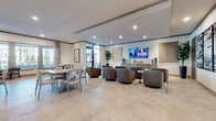 This well-appointed single-floor community clubhouse offers a thoughtfully designed layout ideal for socializing and relaxation. The main lounge area features a generous arrangement of plush swivel barrel chairs grouped around small accent tables, all anchored by a large wall-mounted flat-screen TV. Neutral-toned large-format tile flooring runs throughout, lending a clean and cohesive aesthetic. A dedicated dining area provides multiple tables with mixed seating, bathed in natural light from expansive windows that look out onto an outdoor courtyard. An adjacent corridor-style entry hall showcases a striking geometric parquet-style wood floor, open shelving, and a decorative pendant chandelier, creating an inviting transition between spaces. The full-service kitchen bar is a standout feature, offering an elongated granite-topped island lined with upholstered bar stools, complemented by stainless steel appliances, custom cabinetry, and an eye-catching patterned tile backsplash. A separate game and entertainment room includes a billiards table, a round dining table with modern seating, wall-mounted TV, and sputnik-style chandeliers. Throughout the space, curated artwork, tall indoor plants, and wall sconces contribute to a polished, contemporary ambiance designed for both casual gatherings and community events.