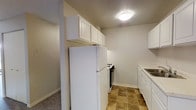 This single-floor space includes a kitchen designed for efficient use of the available layout. The kitchen features white upper and lower cabinetry that provides ample storage, complemented by a clean, neutral color palette on the walls and ceiling. Essential appliances are in place, including a full-size white refrigerator, a gas range with black finish, and a built-in sink with surrounding counter space. The flooring in the kitchen consists of earthy-toned square tiles, offering durability and easy maintenance. A flush-mount ceiling light provides consistent overhead illumination throughout the kitchen area. A structural column is incorporated into the kitchen layout, adding a distinctive architectural element while housing additional upper cabinetry above the refrigerator. Adjacent to the kitchen, a section of carpeted flooring is visible, suggesting a transition into a living or hallway area. The overall aesthetic is straightforward and practical, utilizing light tones to create a clean, open feel within the space.