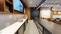 This single-floor space showcases a thoughtfully designed interior that blends functionality with modern aesthetics. The kitchenette area features a clean white countertop with an undermount sink, complemented by sleek cabinetry with brushed metal hardware. A wall-mounted flat-screen TV is integrated above the cabinetry, framed by warm wood paneling that adds a rich, natural contrast to the otherwise cool-toned palette. A geometric patterned floor in muted grey and white tones runs the length of the space, adding visual texture underfoot. Recessed and track lighting illuminate the area efficiently, highlighting the refined finishes throughout. Adjacent to the kitchenette, a long dark-toned communal table is flanked by tan upholstered chairs, creating a defined dining or meeting area. A deep navy blue accent wall anchors the far end of the space, while a textured white architectural panel on the side wall adds dimension and visual interest. The overall ambiance is polished and professional, making this space well-suited for gatherings, meetings, or collaborative events.
