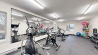 This single-floor fitness facility offers a thoughtfully designed space for a comprehensive workout experience. The main exercise room is generously proportioned, featuring dark, durable flooring that provides a practical and clean aesthetic throughout. Bright fluorescent lighting illuminates the entire area evenly, ensuring excellent visibility during any activity.

The exercise room is equipped with a diverse selection of fitness equipment, including spin bikes, an elliptical trainer, a treadmill, and a rowing machine, catering to a variety of cardio preferences. A dedicated free weights area houses a full rack of dumbbells and plate-loaded equipment, while a medicine ball rack adds versatility for functional training. A large wall-mounted mirror runs along one side of the room, enhancing the sense of openness and allowing users to monitor their form. A wall-mounted mini-split air conditioning unit ensures climate comfort year-round, and windows along one wall invite natural light and outdoor views.

The facility also includes a well-appointed, accessible bathroom. Clean white walls, neutral tile flooring, a floating wood-accented vanity with mirror, and stainless steel grab bars reflect a practical yet modern design. A paper towel dispenser and additional fixtures underscore the space's commitment to functionality and accessibility.