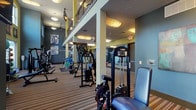 This two-floor fitness center is thoughtfully designed to support a comprehensive range of workout routines. The space is finished with striped carpet flooring throughout and features a color palette of muted blues, greens, and yellows that lends the facility a modern, energetic feel. Structural columns and open railings define the split-level layout, with a staircase connecting the two floors.

The upper level is dedicated primarily to cardio training and is lined with an extensive selection of equipment, including treadmills, elliptical machines, and stationary cycling bikes. Ceiling-mounted televisions are positioned throughout this level for convenience. Large windows along the perimeter flood the floor with natural light and offer views of the surrounding outdoor greenery. The stairwell area also features floor-to-ceiling windows that further enhance the sense of openness.

The lower level serves as the strength training hub of the facility. It houses a variety of cable and selectorized machines, squat racks with Olympic barbells and weight plates, benches, and additional specialty equipment. Full-length mirrors line key sections of the walls, maximizing the sense of space and supporting proper form during workouts. The lower level also includes a designated free-weight area with rubber flooring mats. Safety features such as a fire extinguisher and posted fitness center rules are clearly visible throughout the space.