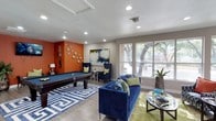 This single-floor community amenity space is thoughtfully designed to offer residents and visitors a welcoming, multi-functional environment. The interior is characterized by warm hardwood-style flooring, recessed lighting, and an upbeat color palette that combines bold orange accent walls with navy, green, and neutral tones throughout.

The social lounge is a standout area, anchored by a full-size pool table set atop a bold geometric-patterned area rug. A mounted flat-screen TV, wall-mounted cue rack, and a mix of plush navy sofas and wingback chairs create an inviting entertainment atmosphere. Decorative starburst wall accents and a large abstract painting add artistic flair to the space.

An adjacent seating area transitions smoothly into a bright workspace or leasing office, lined with oversized windows that flood the room with natural light and offer views of mature trees outside. Patterned accent chairs with orange throw pillows, a bookcase, and a dedicated desk area make this zone both functional and comfortable.

A clean, well-appointed kitchenette with white cabinetry, pendant lighting, and a wide sink provides practical convenience, while a built-in water fountain nearby further enhances the facility's amenities. The open, flowing layout connects each area seamlessly, making the overall space feel expansive, lively, and purpose-built for community engagement.