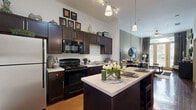 This 1-bedroom, 1-bathroom residence is laid out entirely on a single floor, making smart use of its cozy footprint. The open-concept kitchen and living room serves as the heart of the home, featuring rich dark-espresso cabinetry extending to the upper walls, light-toned countertops, and a central island with pendant lighting overhead — ideal for both casual dining and meal prep. Black appliances, including a range, built-in microwave, and stainless steel refrigerator, complement the kitchen's contemporary aesthetic. Warm hardwood-style flooring runs throughout the kitchen and entry areas, lending a polished yet inviting character to the space. The living room benefits from soaring ceilings and large windows that allow natural light to fill the room, creating an airy, open ambiance. Neutral sage-toned walls provide a calming backdrop throughout. The bedroom is a comfortable retreat with plush carpeting, large windows with outdoor views, and ample room for a full furniture arrangement. Two closets provide generous storage. The bathroom features dark wood vanity cabinetry, a wide mirror, and a full tub-shower combination, finished with tile flooring. A hallway connects all rooms, completing this well-organized and stylish layout.