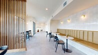 This thoughtfully designed single-floor commercial space offers a bright, welcoming environment defined by a clean, modern aesthetic. The layout is organized into several distinct zones, each with its own character while maintaining a cohesive design language throughout.

At the heart of the space is a bold statement wall clad in white shiplap paneling, bearing the motivational phrase "Dare to Be Bold" in dimensional lettering, flanked by gold accent sconces — setting an energetic tone for visitors. A seating nook nearby features mid-century modern armchairs arranged around a sculptural coffee table atop a woven area rug.

A long banquette seating area commands one wall, upholstered in caramel-toned channeled leather beneath a backdrop of white glazed tile. Marble-topped tulip tables paired with black dining chairs provide stylish seating for multiple guests, illuminated by warm gold wall sconces.

A wooden slatted partition divides the space gracefully, leading to a counter-height bar ledge along a floor-to-ceiling window wall that frames views of lush palm trees and the outdoors, flooded with natural light. Throughout, large-format gray tile flooring ties each area together seamlessly, complemented by recessed ceiling lighting and globe pendant fixtures that enhance the warm, inviting ambiance.