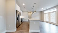 This single-floor unit on Western Centre Drive in Houston, Texas presents a well-designed, modern interior with a cohesive neutral color palette and wood-look flooring that extends throughout the entire space. The open-concept layout connects the kitchen and living area seamlessly, creating a functional and visually spacious environment.

The kitchen is a standout feature, equipped with stainless steel appliances including a French door refrigerator, built-in microwave, and range. Crisp white cabinetry with black hardware provides clean contrast, while granite countertops add a polished finish. A central island with an integrated sink offers additional prep space and seating potential. Three brushed gold pendant lights hang above the island, adding a contemporary design element.

The living area receives natural light through large windows and a set of French doors, which also provide access to the exterior. A ceiling fan with a light fixture services this area, enhancing comfort. The space flows openly from the kitchen, making it ideal for everyday living.

A private bedroom features wood-look flooring, a window with exterior views, and a ceiling-mounted light fixture. The bathroom is finished with white subway tile surrounding the tub-shower combination, a granite-top vanity with white cabinetry and black hardware, and a framed mirror.