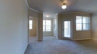 This single-floor residence offers a thoughtfully designed open-concept layout that seamlessly connects the main living areas. The living room is generously sized, featuring plush wall-to-wall carpeting, crown molding, and a ceiling fan with light fixture that adds both comfort and style. Track lighting provides additional illumination, complementing the abundant natural light that streams in through large windows fitted with blinds. A door leads directly to a private outdoor area, enhancing the sense of openness and accessibility.

The kitchen is partially open to the living space, separated by a structural column and a peninsula counter finished with granite-look countertops and a dark base — a functional and attractive design element. A sink is visible at the edge of the counter, and a hallway beyond leads to additional rooms.

A secondary carpeted room, visible through an open doorway, mirrors the living room's warm, neutral color palette and also benefits from natural light through its own window. Crown molding carries throughout, providing a cohesive and polished finish to the interiors. The overall ambiance is clean, neutral, and move-in ready, with warm beige tones on the walls and crisp white trim creating an inviting and comfortable atmosphere throughout.