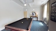 This dedicated game room offers a well-appointed space designed for recreation and social gatherings. The centerpiece of the room is a full-size billiards table with a sleek dark felt surface, positioned beneath a striking coffered ceiling that adds architectural depth and a refined aesthetic to the space. Elegant pendant lights hang overhead, providing focused illumination while complementing the room's polished design sensibility. Crown molding frames the upper walls, lending a classic, finished look throughout. The floor is covered in a textured blue-toned carpet that adds warmth and a contemporary feel underfoot. Along the wall, a wooden console table serves as a decorative accent, styled with table lamps and a bold geode-inspired artwork that introduces a pop of color and artistic flair. A curtained window allows natural light to filter into the space, brightening the neutral, light-gray walls. A private door provides easy access to adjacent areas, ensuring convenience without interrupting the room's cohesive layout. Overall, the game room strikes a balance between functional recreation and thoughtful interior design, making it an inviting destination for leisure and entertainment.