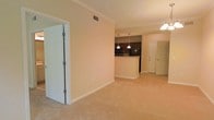 This single-floor residence presents an open-concept layout that seamlessly connects the main living area to the kitchen, creating a cohesive and functional flow. The living space is finished with plush, light-toned carpeting underfoot and features warm beige walls complemented by crisp white crown molding and baseboards, lending a clean, polished appearance throughout.

The kitchen is positioned at the rear of the open living area and is defined by a raised peninsula that provides both a workspace and a natural dividing element between the two zones. Dark cabinetry with rich wood tones contrasts against the lighter surroundings, while black appliances, including a refrigerator and microwave, contribute to a cohesive, contemporary kitchen aesthetic. Pendant lighting hangs above the peninsula, offering focused task lighting and an added design detail.

A ceiling-mounted light fixture in the living area provides general illumination, while a ceiling vent indicates a central HVAC system. Panel-style doors are visible, including what appears to be a closet and an entry or utility door. The overall ambiance is neat, neutral, and move-in ready, offering a comfortable and straightforward living environment.