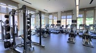 This fitness center is thoughtfully designed to support a comprehensive workout experience. The space is bathed in natural light, courtesy of large windows framed in warm wood trim that line the walls, creating an open and energizing atmosphere. The high ceilings contribute to the spacious feel, ensuring the room never feels crowded even when fully equipped.

The cardio area is well-stocked with a row of treadmills, elliptical trainers, and a stair climber, all positioned to take advantage of the natural light and outdoor views. Multiple flat-screen televisions are mounted at convenient viewing angles throughout the space, allowing users to stay entertained during their workouts.

On the strength training side, a robust power rack with an assortment of weight plates anchors the free weights area. Full-length mirrors are strategically placed along one wall, enhancing the sense of space and allowing users to monitor their form during exercises.

The flooring throughout is durable rubber matting, providing both safety and comfort underfoot. The overall ambiance is clean, modern, and functional, making this fitness center a well-rounded facility suitable for a wide range of fitness routines.