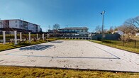 Located at 346 W 26th St in Charlotte, NC, this outdoor amenity space is designed to serve as a recreational hub within a developing residential community. The centerpiece of the area is a full-size sand volleyball court, complete with a regulation net and boundary markings. A dedicated light pole positioned courtside ensures the court remains usable during evening hours, making it a functional year-round amenity. The surrounding grounds are well-maintained, featuring neatly trimmed grass borders that frame the court and adjacent hardscaped areas. Wide concrete walkways connect the various sections of the site, providing accessible circulation throughout. A gated entry point with wooden panel accents and iron fencing adds a layer of security and visual structure to the perimeter. Nearby, utility infrastructure is discreetly integrated into the landscape. The broader setting includes a multi-story residential building with white siding, brick detailing, and private balconies with wood-accented overhangs, visible in the background. Additional construction activity in the surrounding area indicates continued neighborhood growth. The open-air design, abundant natural light, and spacious layout create an inviting environment well-suited for active outdoor recreation.